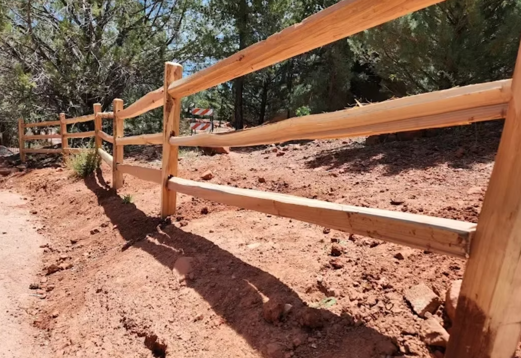 Wooden fence with orange mesh lining a dirt trail in a sunny wooded area