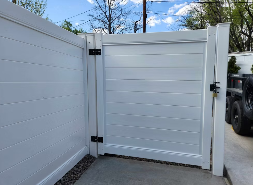 White slatted metal gate with black hinges and latch beside a driveway wall.