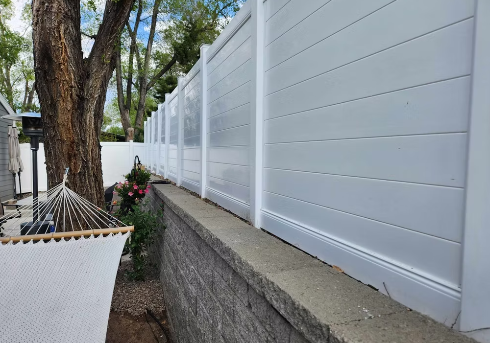 White privacy fence along a stone wall beside trees and a rope hammock on a patio