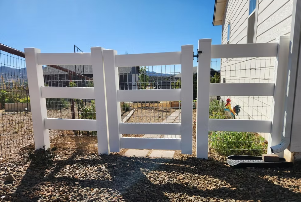 White fence gate in a gravel yard with desert plants and a house wall on the right.