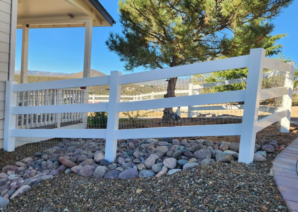 White wooden fence beside a covered patio, with rocks in front and a pine tree behind.