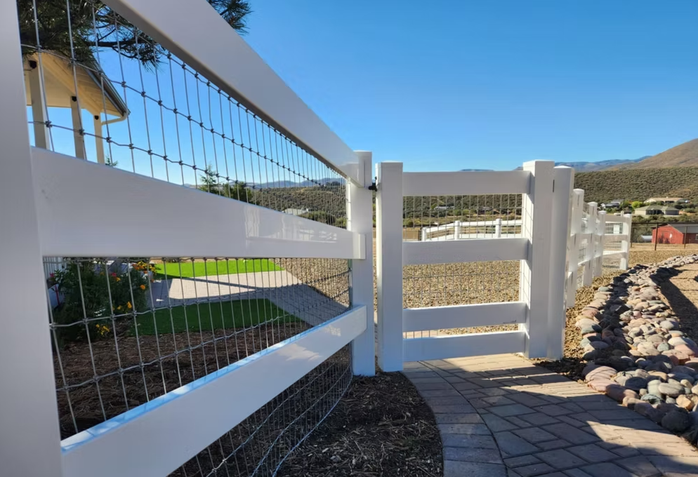 White ranch fence with wire mesh and gate beside a paved path in a sunny rural area