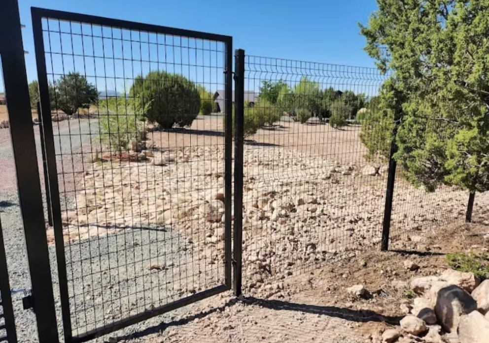 Open black metal gate in a desert yard with shrubs and a paved path under a clear blue sky