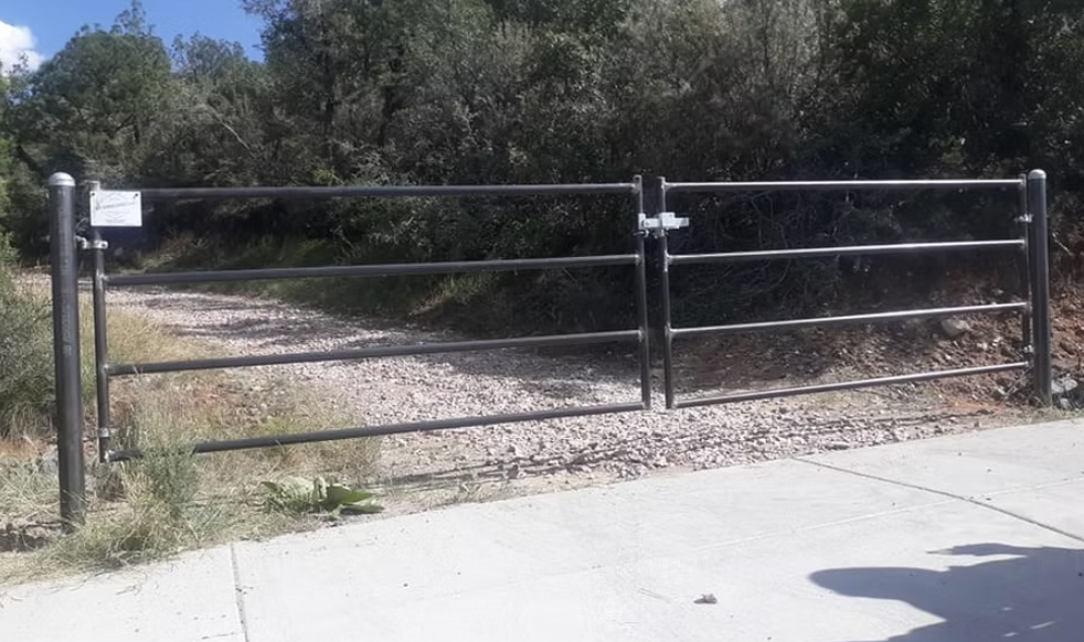 Closed metal gate blocking a gravel trail beside a concrete road, with a small sign on the left.
