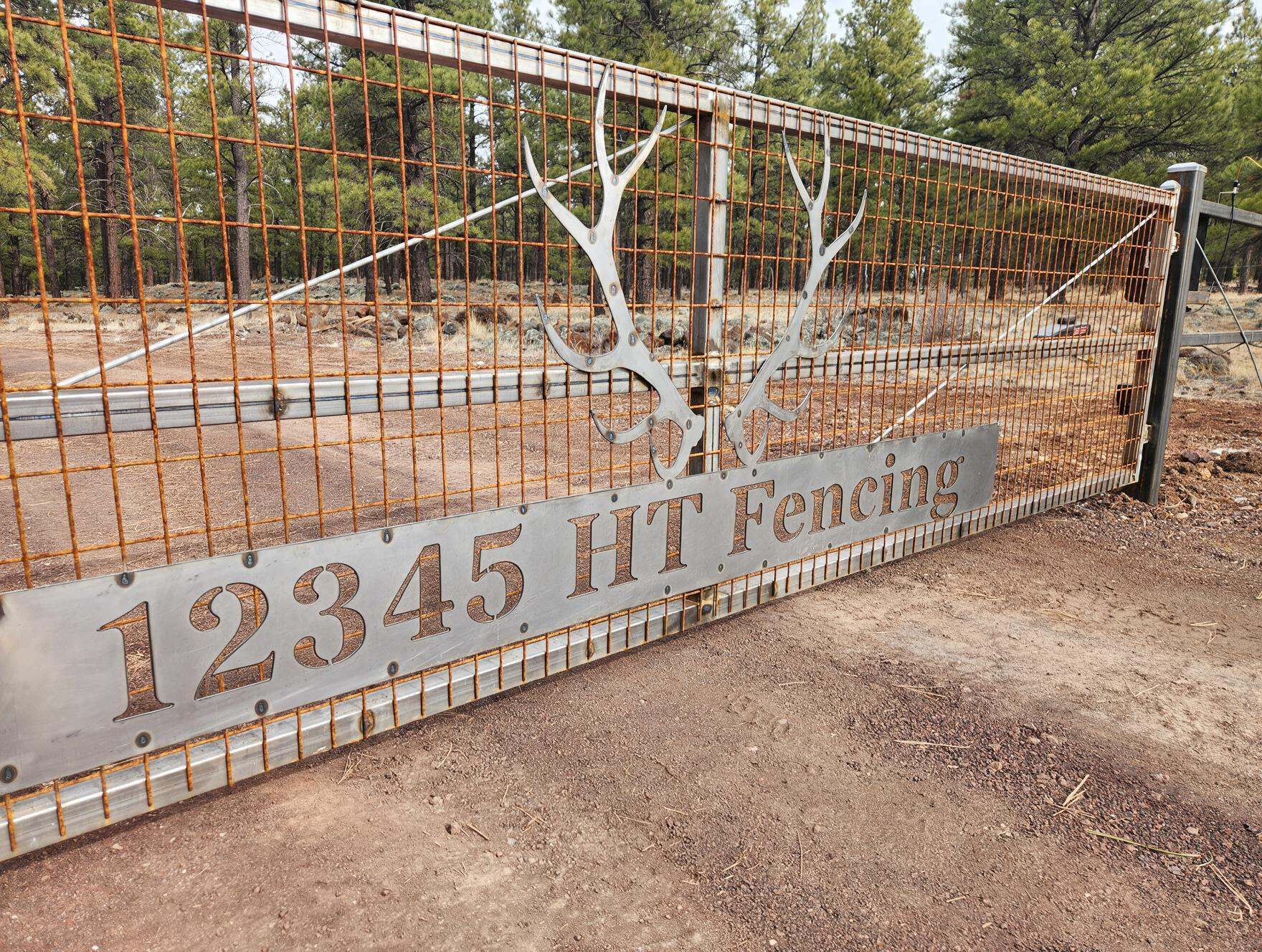 Rust-colored metal gate with “12345 HT Fence” sign in a gravel driveway, trees behind it