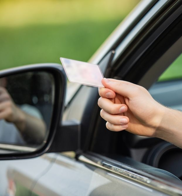 Hand holding driver's license out of car window, with side mirror and green background visible.