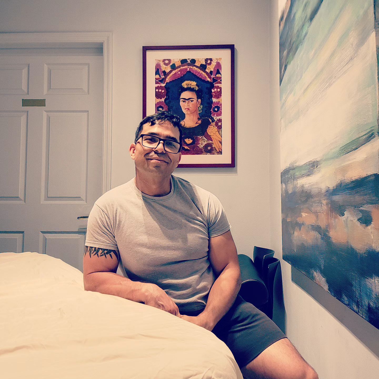 Man in glasses, gray shirt, and shorts sits on a bed, smiling; Frida Kahlo portrait and abstract painting on wall.