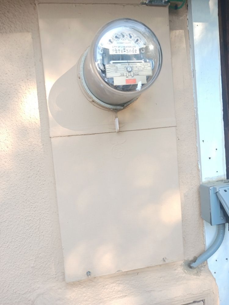 Residential electric meter mounted on a beige exterior wall with conduit and utility box