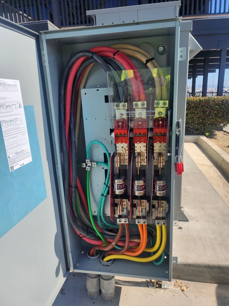 Open electrical panel with exposed breakers, red and black cables, and green grounding wire outdoors.