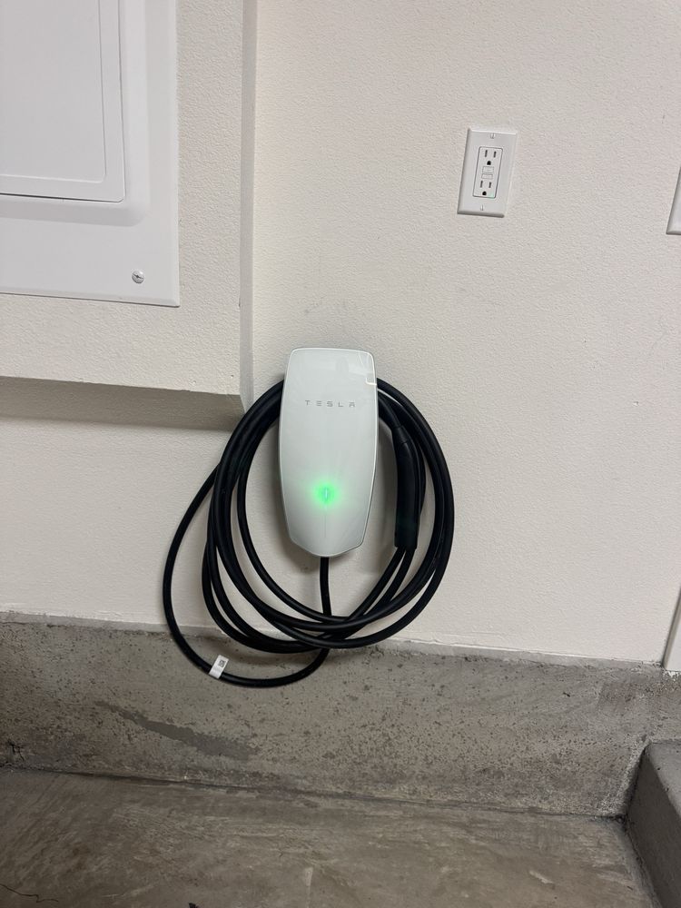 Wall-mounted EV charger with a glowing green light and coiled black cable next to an outlet.