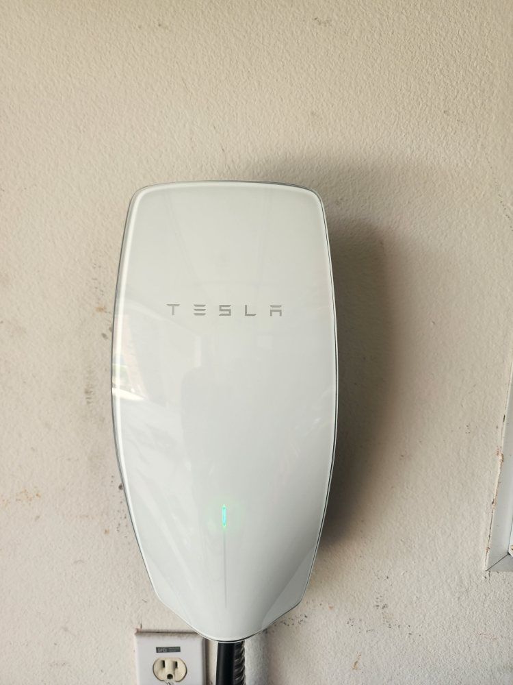 White Tesla wall-mounted EV charger on a beige wall above an electrical outlet.