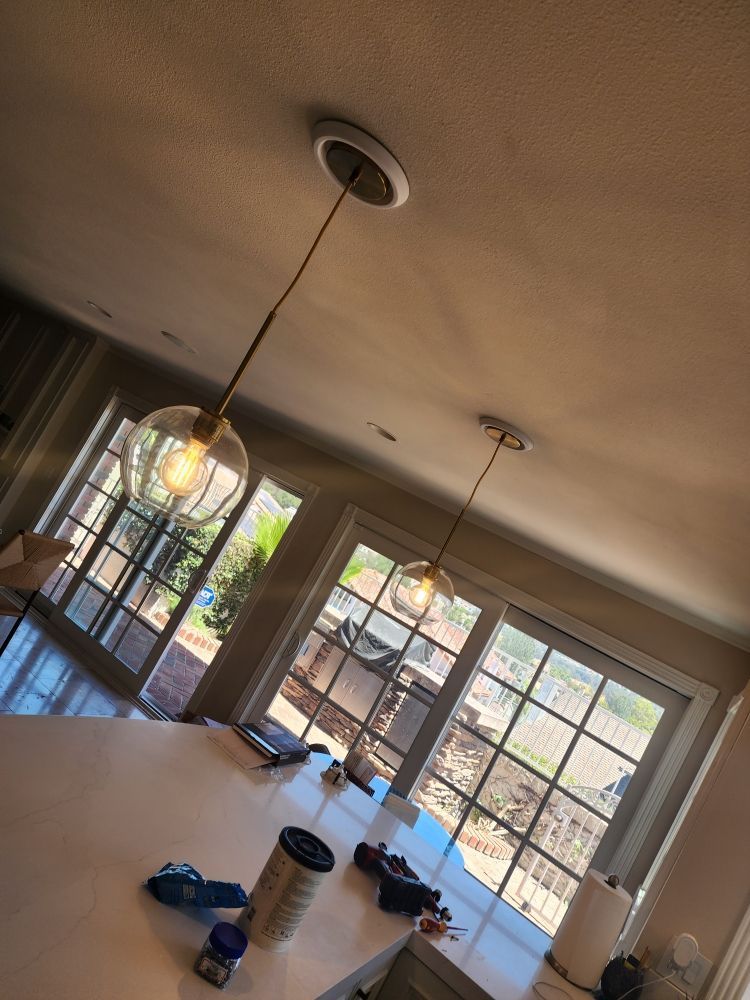Sunlit living room with large windows, pendant lights, and scattered items on a table and floor.