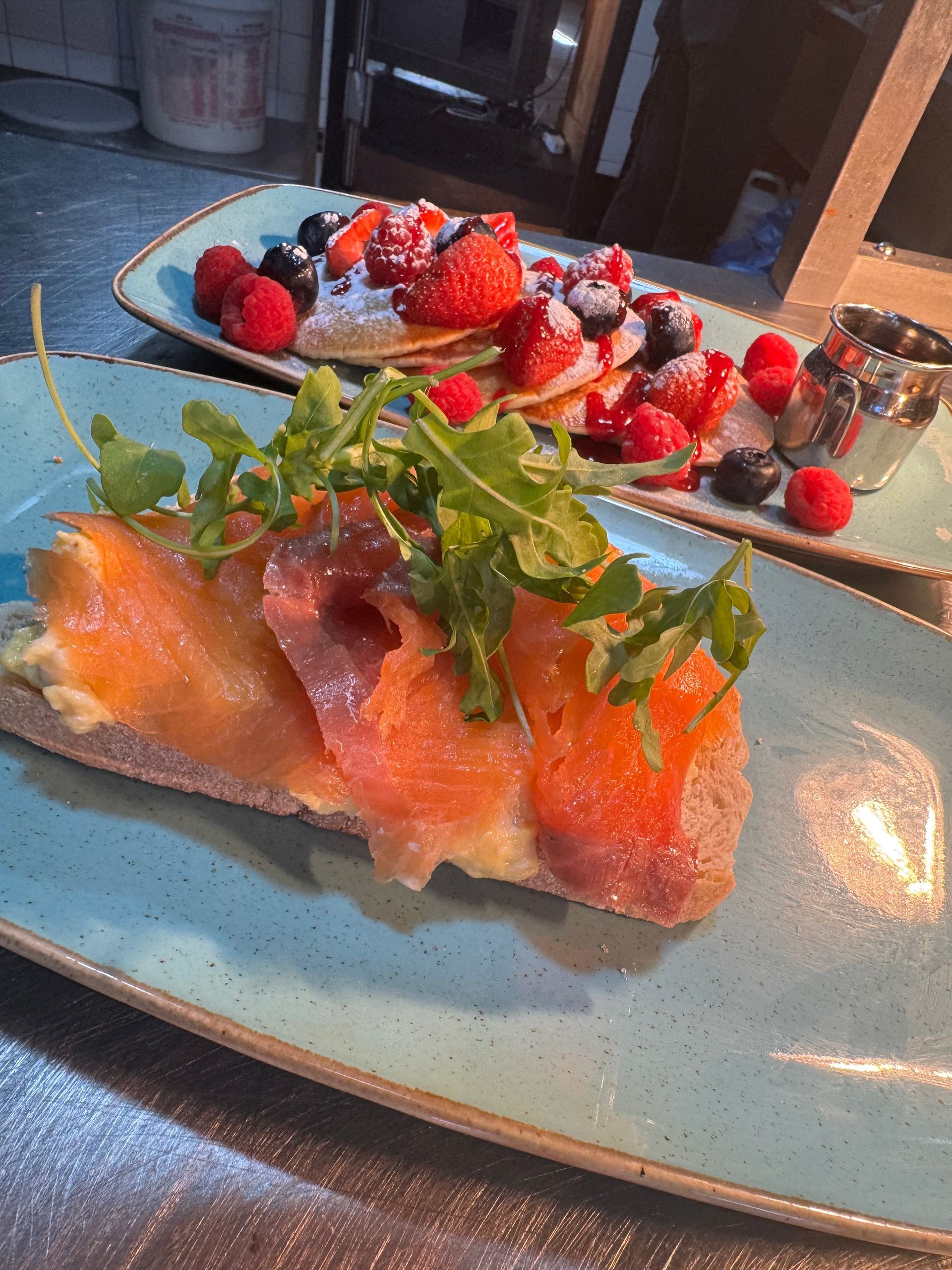 Plated salmon with greens in the foreground and a berry dessert in the background on a table