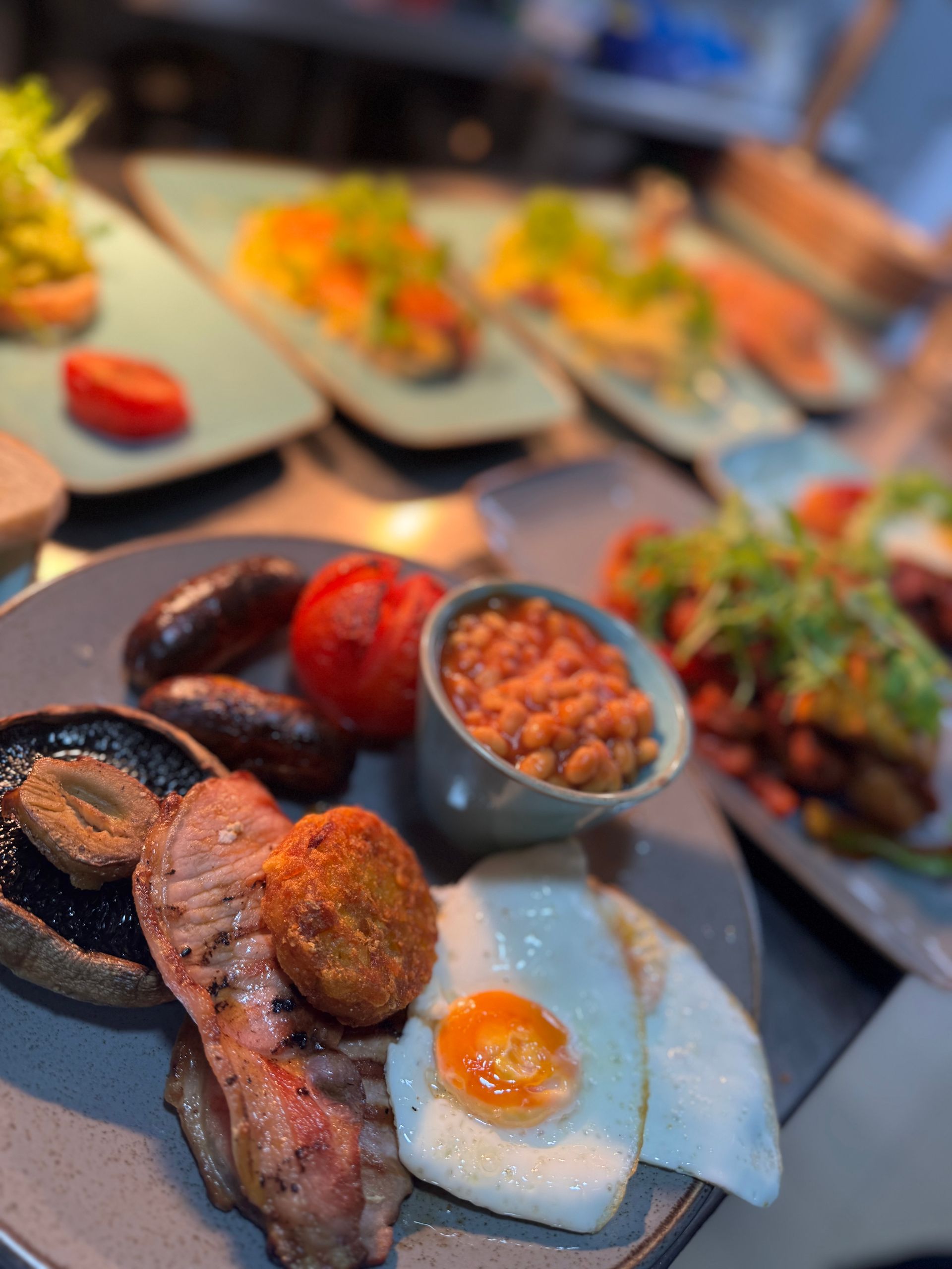 Plated breakfast with sausage, beans, fried egg, and toast on a table with other dishes in the background