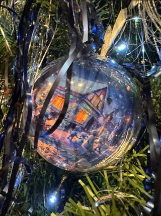 A glass Christmas ornament with a painted house hangs among evergreen branches and lights.