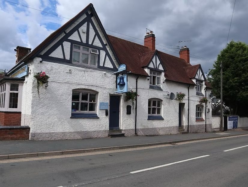 White half-timbered cottage along a road under cloudy skies, with red roof and hanging flower baskets