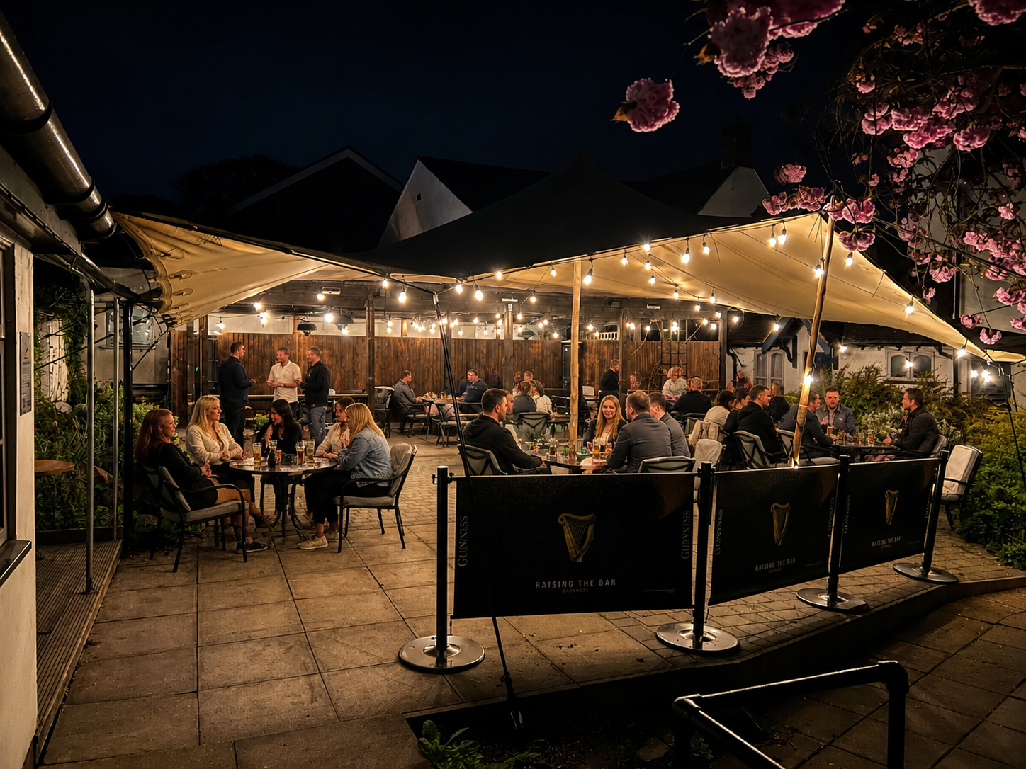 Outdoor café patio at night with warm string lights, seated diners, and pink flowering trees in the background