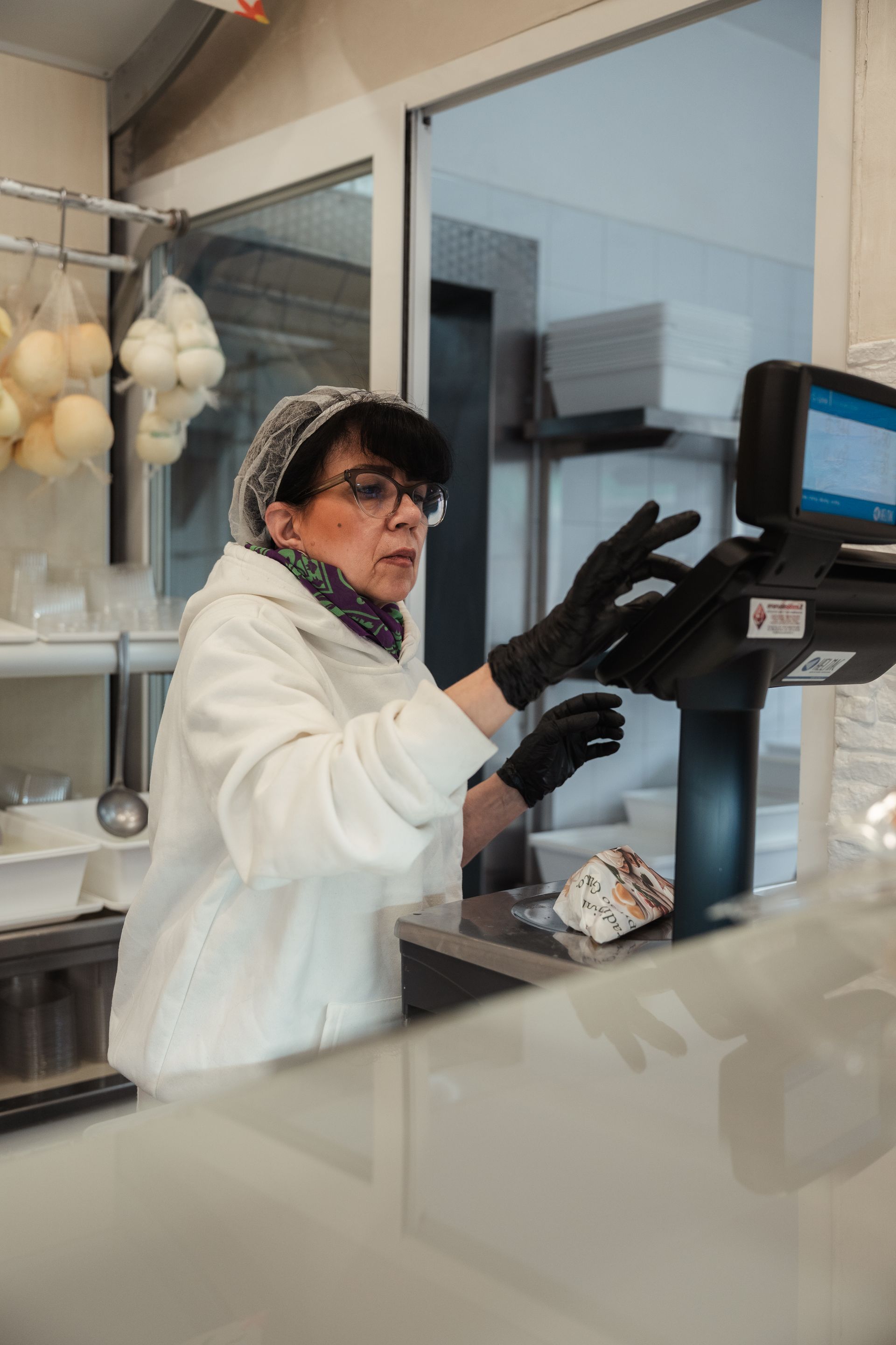 Un operaio con i guanti e il camice bianco utilizza un chiosco touchscreen dietro un bancone in un negozio o bar luminoso.