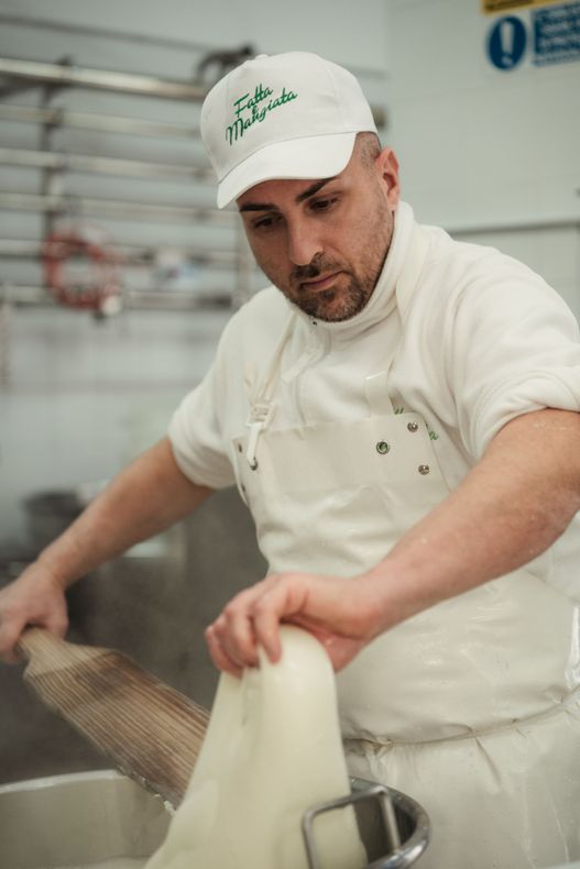 Uno chef in uniforme bianca versa del liquido in una pentola in una cucina professionale.
