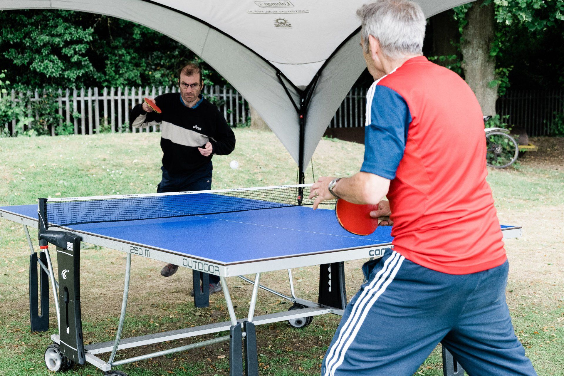 Two men are playing ping pong outside under a tent.