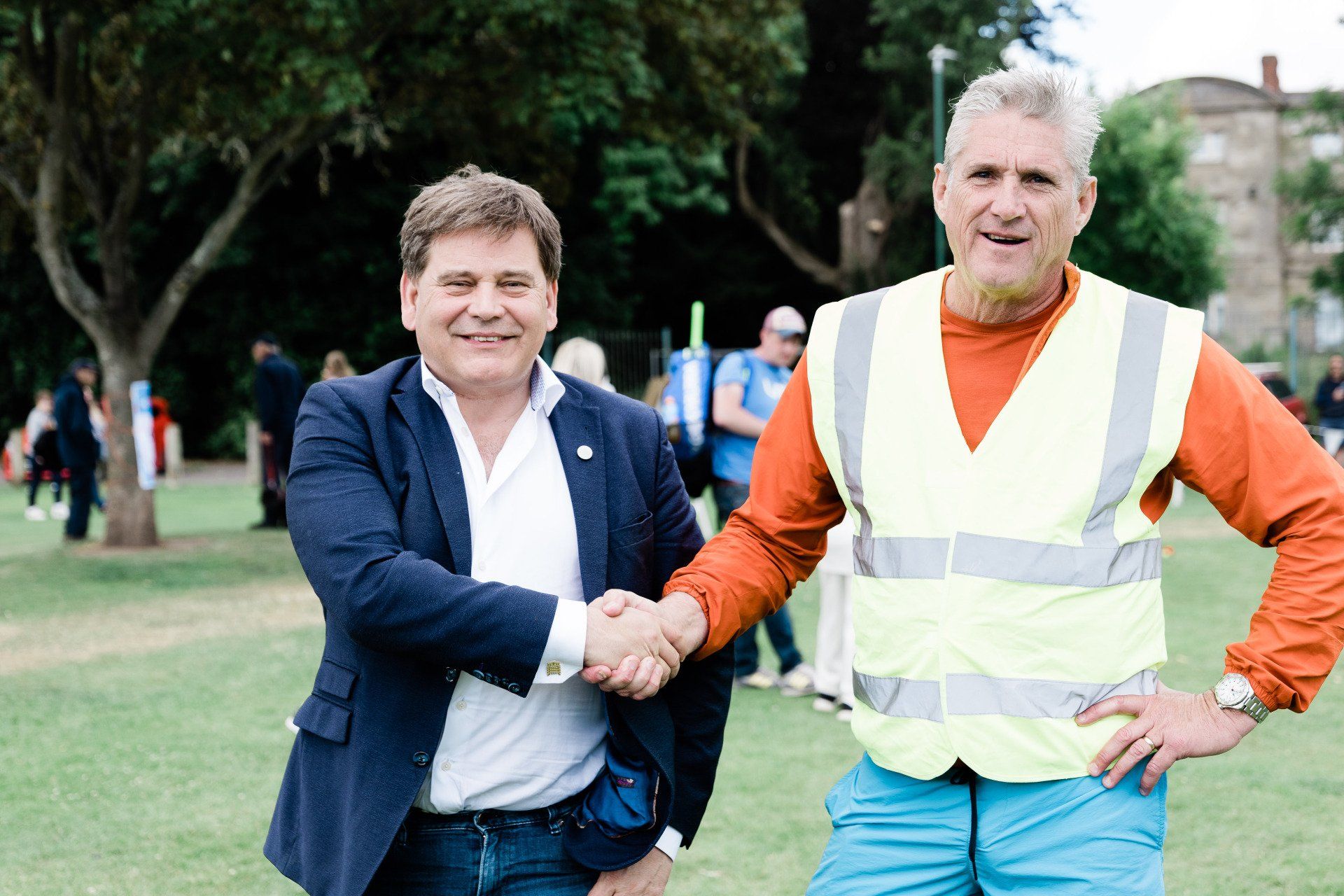 Two men are shaking hands in a park while wearing safety vests.