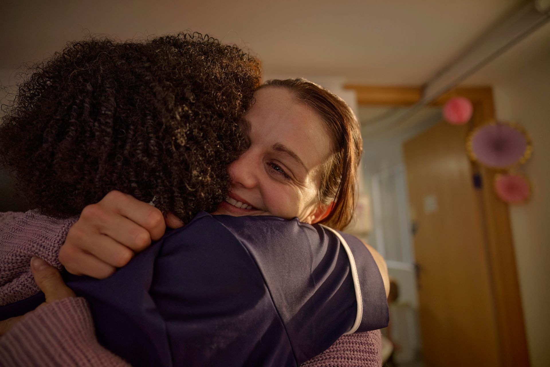 Two people hugging warmly indoors in soft evening light, with a doorway and wall decorations in the background.