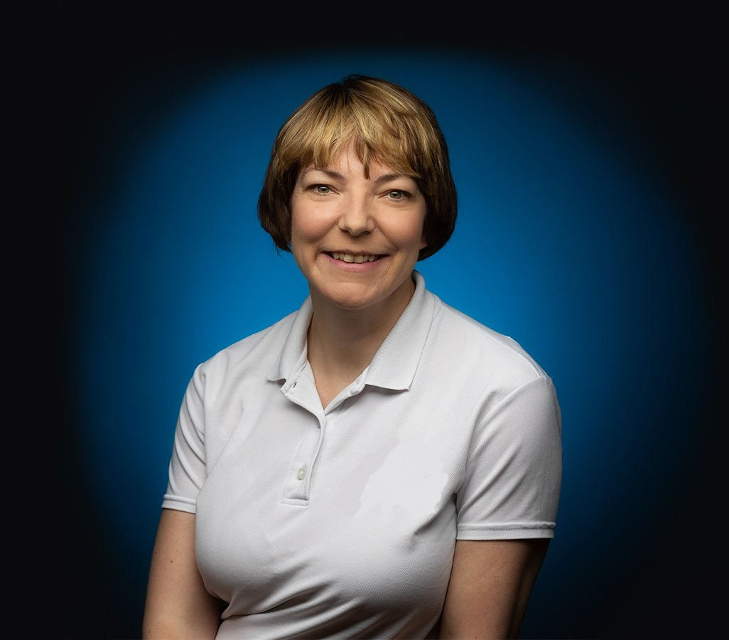 Sharon Betterton, Sports Massage Therapist in a white polo shirt is smiling in front of a blue background.
