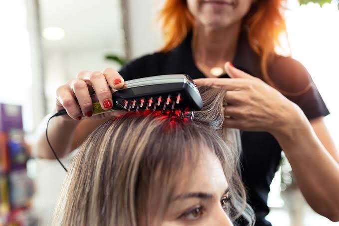 A stylist using a red light therapy brush on a woman's scalp.