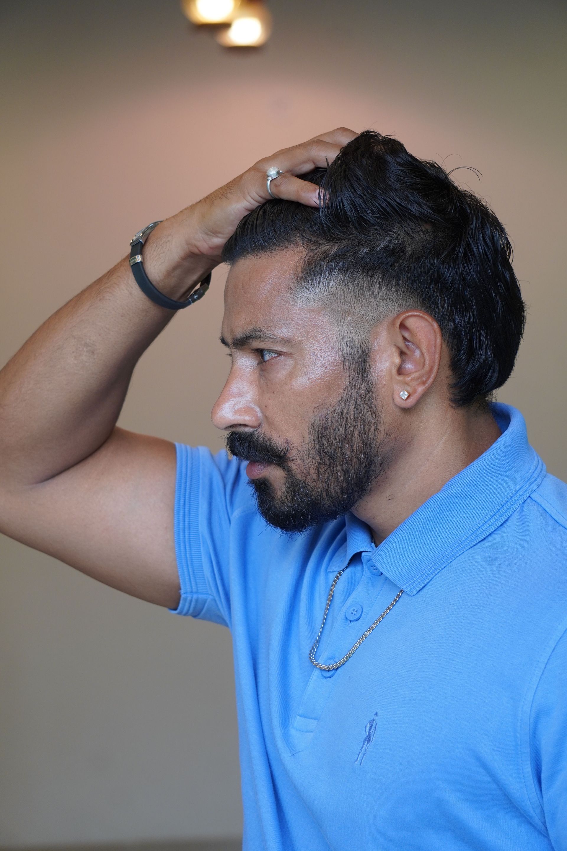 Man in a blue polo shirt touching his hair, shown in profile against a blurred indoor background