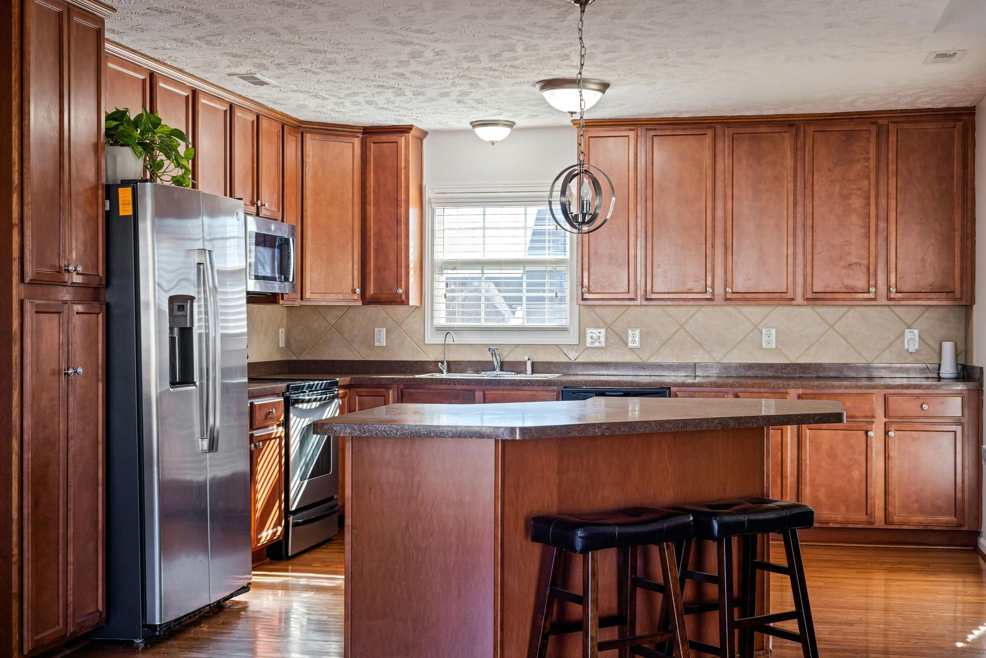 Spacious kitchen with wood cabinets, stainless fridge, island, and two stools under hanging lights.