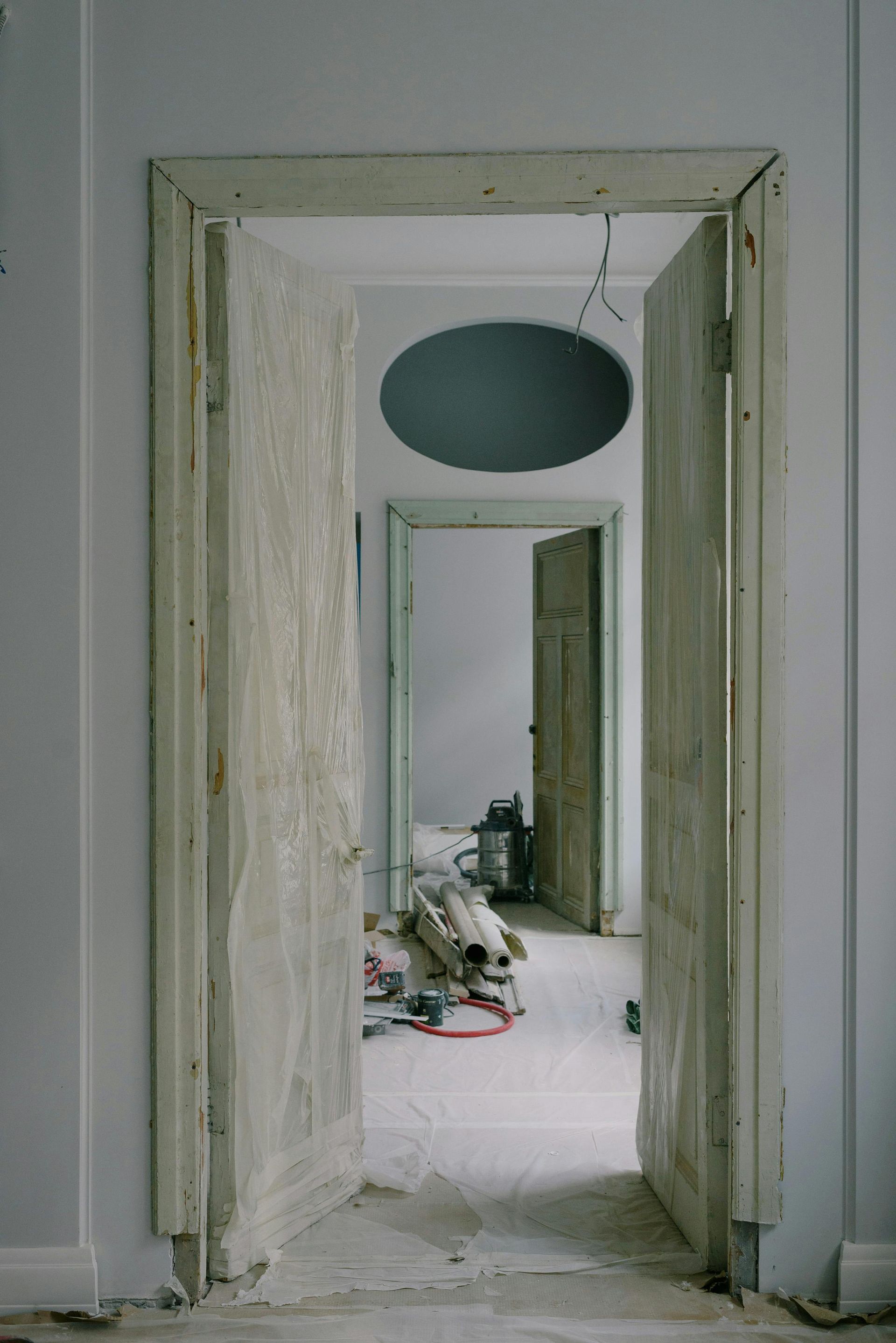 Narrow hallway with peeling walls and a hanging oval light fixture, leading to a cluttered room.
