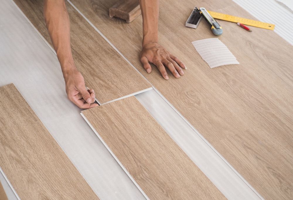 Hands installing light wood vinyl flooring with tools nearby