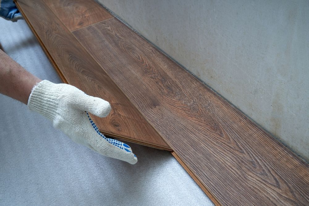 Gloved hand lifting a wood-look plank from a floor underlayment near a wall