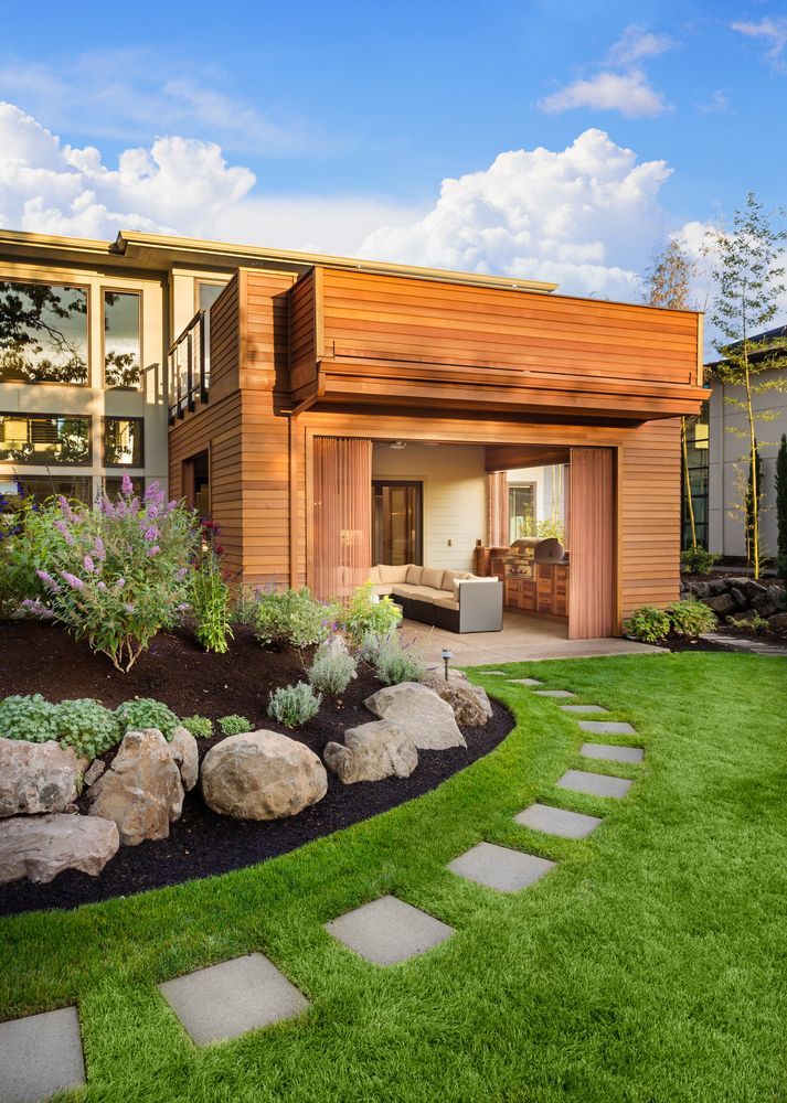 Modern house with a wooden exterior, patio seating, and a curved stepping-stone path through a green lawn.