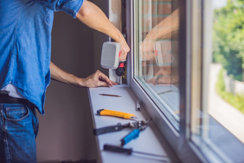 Person using a drill to install a window frame, with tools on the sill.