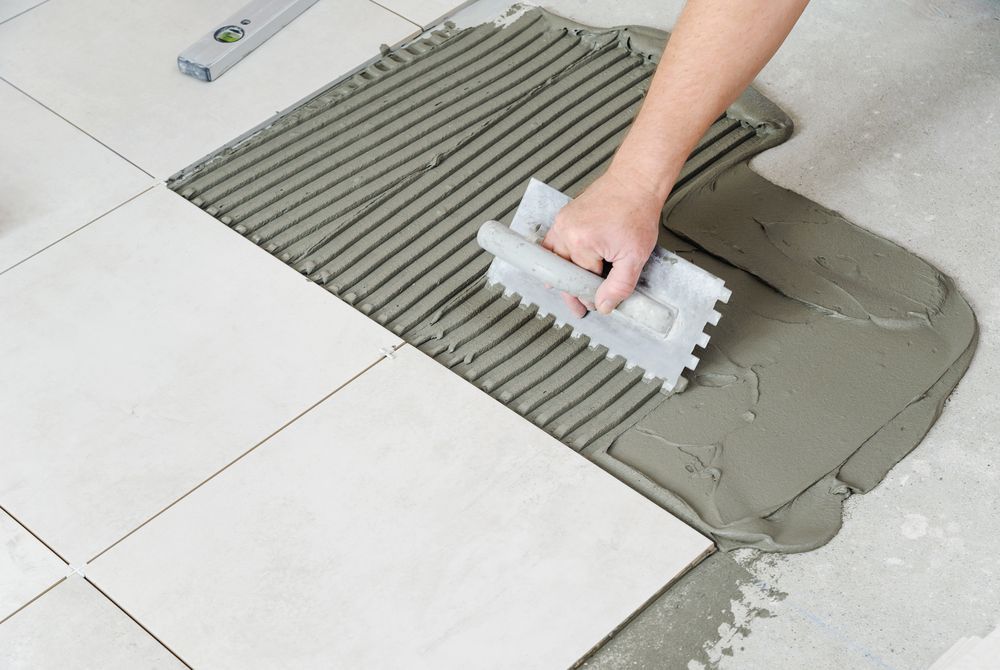 Person spreading gray tile adhesive on a floor with a notched trowel next to white tiles