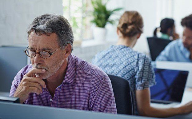 Man in purple shirt and glasses thinking at a laptop in an office, with coworkers blurred in the background