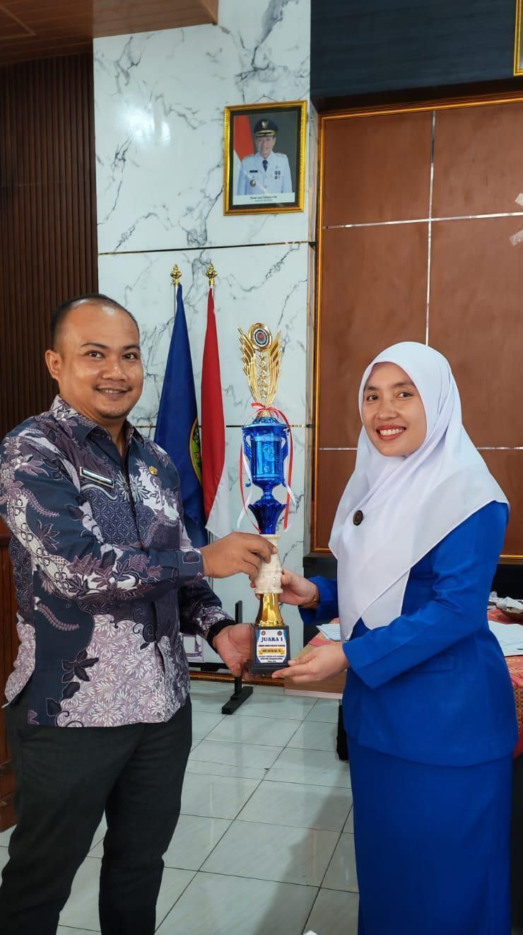 Two people in formal attire holding a trophy in an office with trophies and flags behind them.