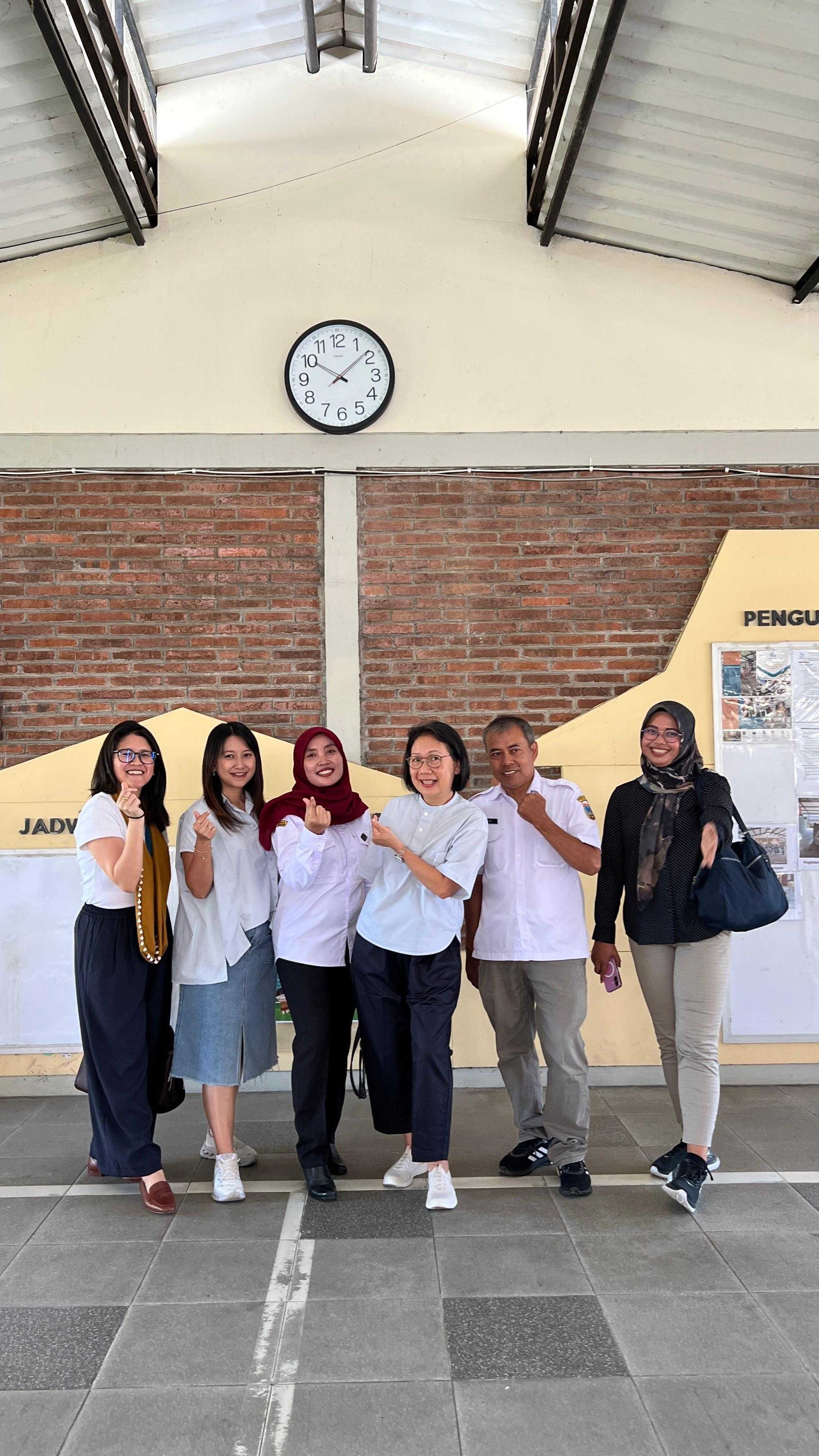 Group of seven people smiling and gesturing in front of a brick wall with a clock.