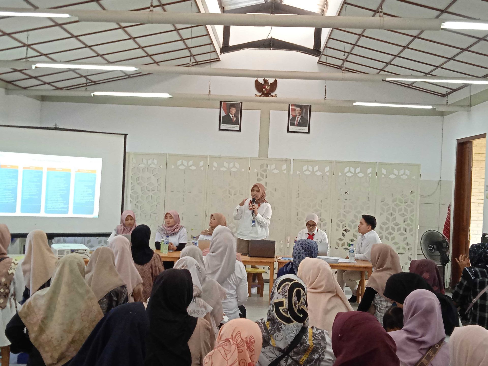 People in a meeting, with a speaker at a podium, presenting slides. Room with Indonesian flags and portraits.