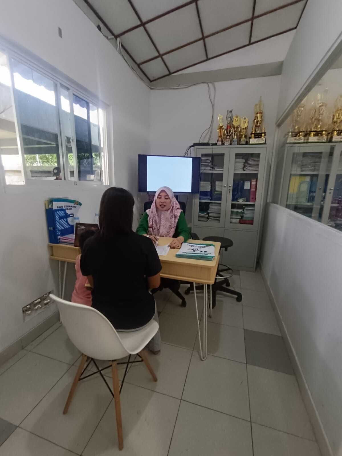 Woman in hijab seated at desk, talking with two others in office setting. Cabinets, screen, trophies in view.