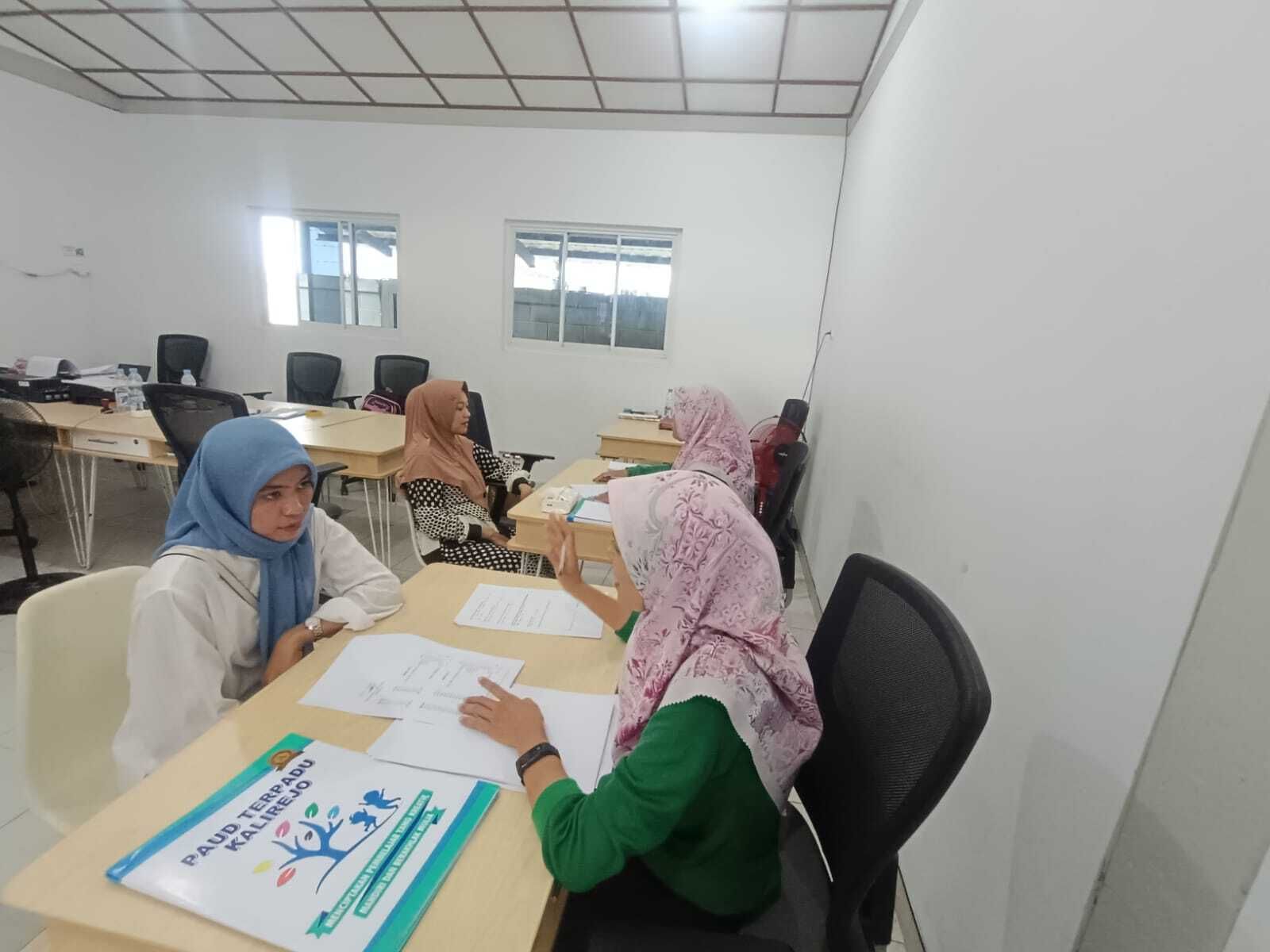 People in headscarves at desks, discussing papers in a classroom.