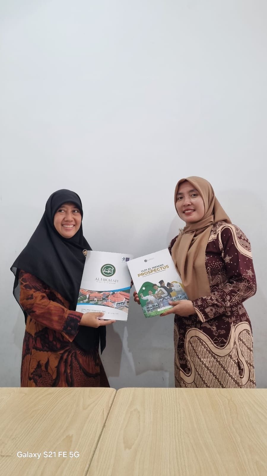 Two people in patterned clothes hold boxed products and smile in front of a plain wall.