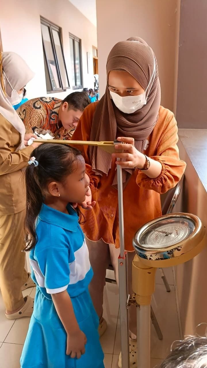 Masked health worker measuring a child’s height in a hallway while others wait nearby