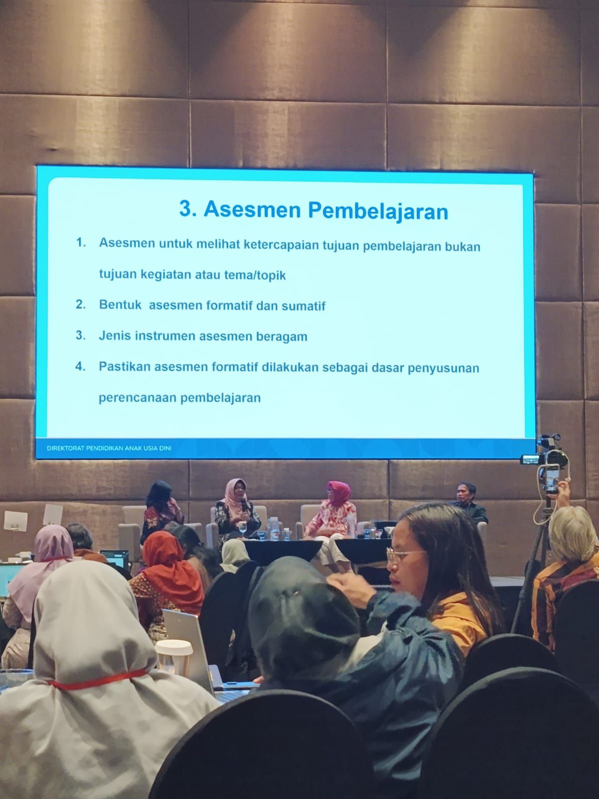 Lecture hall with audience and a blue slide titled “3. Assesmen Pembelajaran” on a projector screen