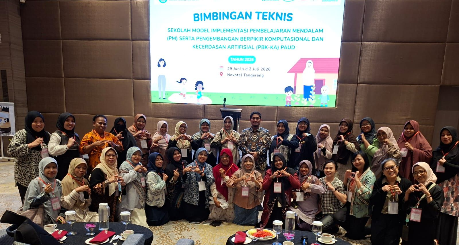 Group photo of people holding drinks at a “BIMBINGAN TEKNIS” event in a conference hall.