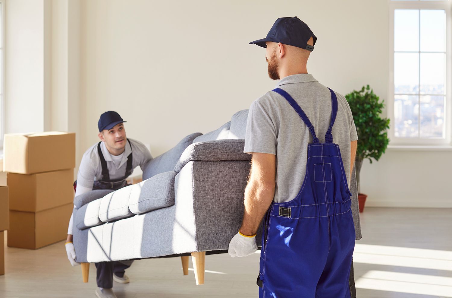 Two professional apartment movers in blue overalls are carefully lifting a gray sofa.