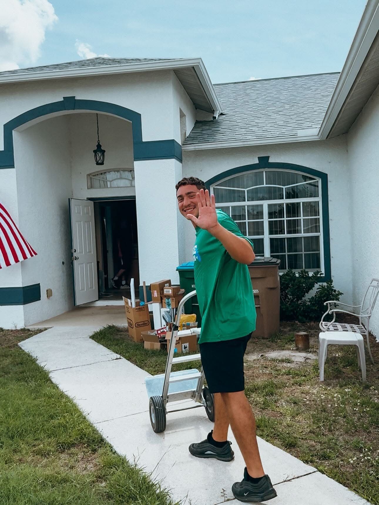 Person waving beside a white house with green trim and a walkway leading to the front door