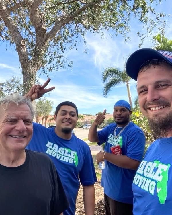Four movers in blue Eagle Moving shirts smiling outdoors by a tree and palm tree.