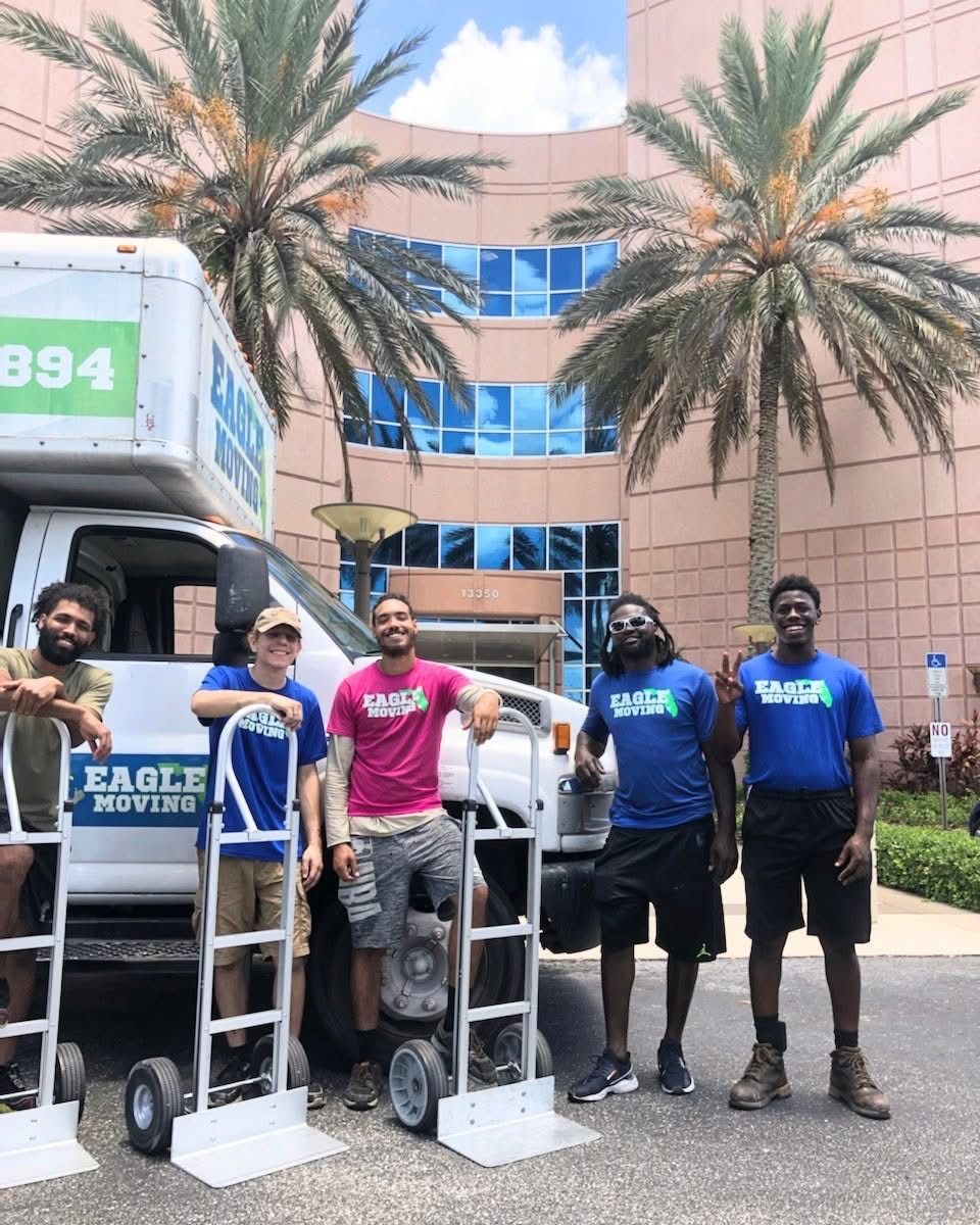Five workers posing beside a truck with equipment carts in front of a building with palm trees